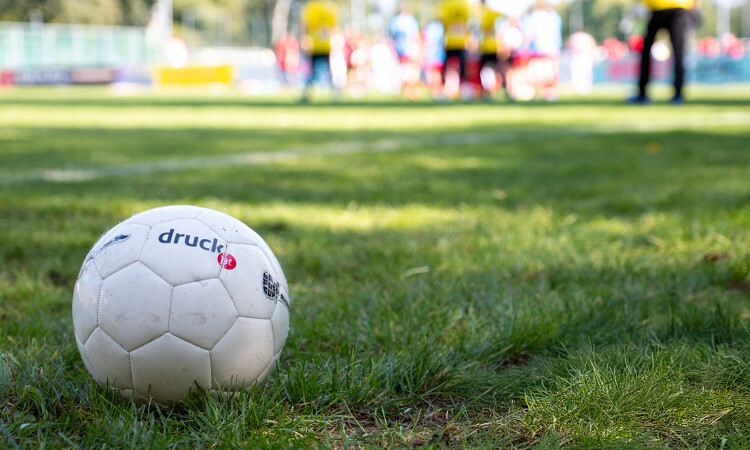 Fußball liegt auf dem Gras, im Hintergrund sind Spieler:innen zu erkennen Fußball liegt auf dem Gras, im Hintergrund sind Spieler:innen zu erkennen
