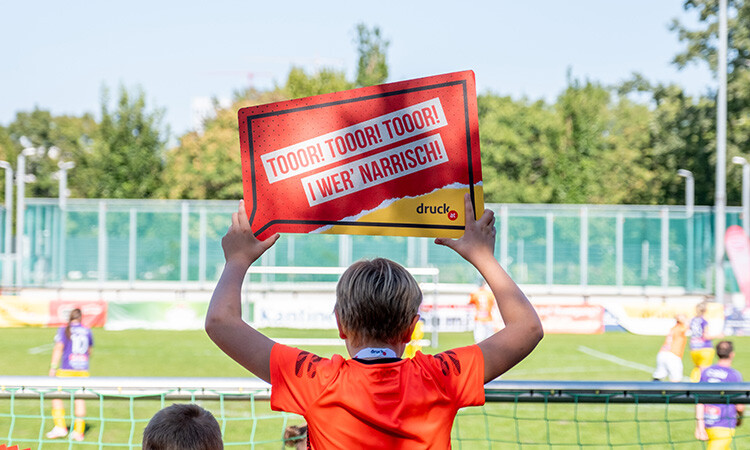 Ein Junge feuert die Spieler:innen mit einem gebrandeten Anfeuerungsschild an Ein Junge feuert die Spieler:innen mit einem gebrandeten Anfeuerungsschild an