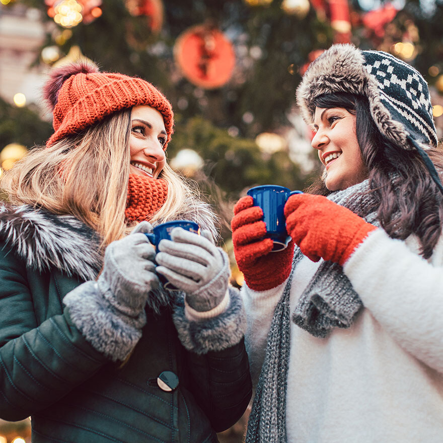 Zwei Personen trinken Punsch vor einem Weihnachtsbaum und lächeln sich an