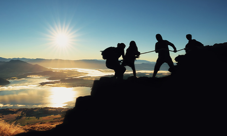 Vier Personen besteigen gemeinsam einen Berg. Im Hintergrund ist ein Sonnenaufgang zu sehen.