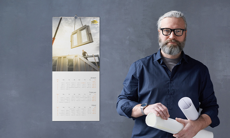 Ein Mann mit schwarzer Brille und grauem Bart hält zwei Papierrollen in der Hand. Er steht neben einem Wandkalender.